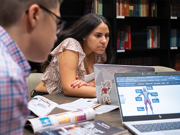 Two students pour through magazines scattered on a table in a library during a research project, with open laptops nearby.